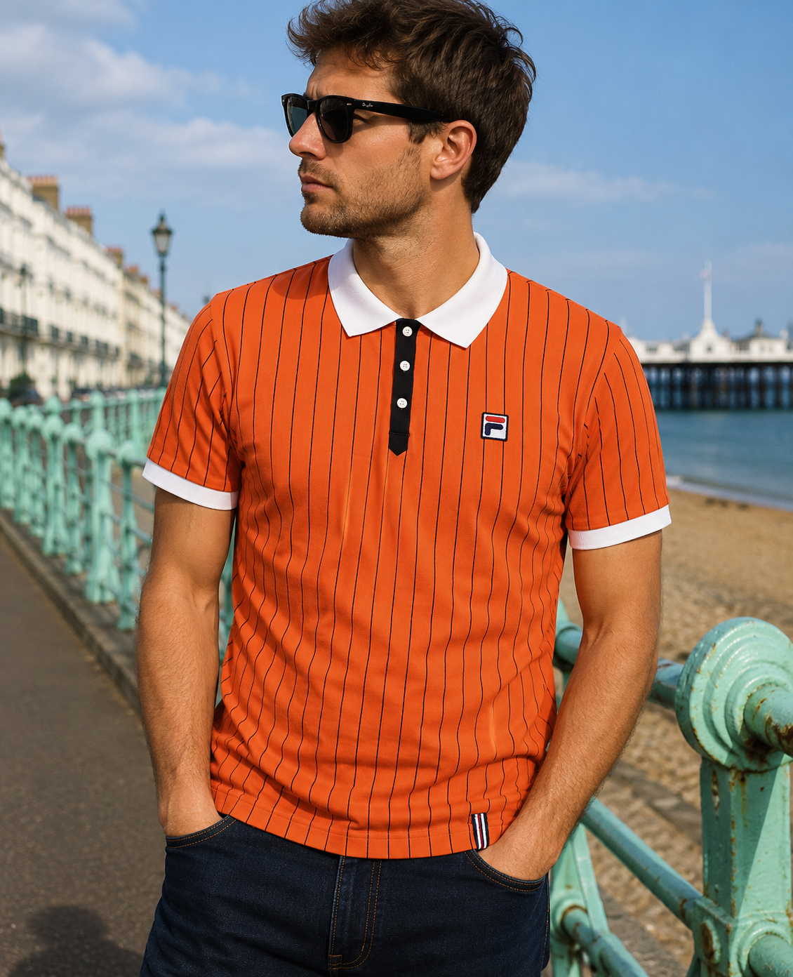 A man wearing an orange short-sleeve polo shirt with thin black pinstripes, a white collar, and a white button placket, standing on a seaside promenade. The shirt features a small Fila logo on the left chest.