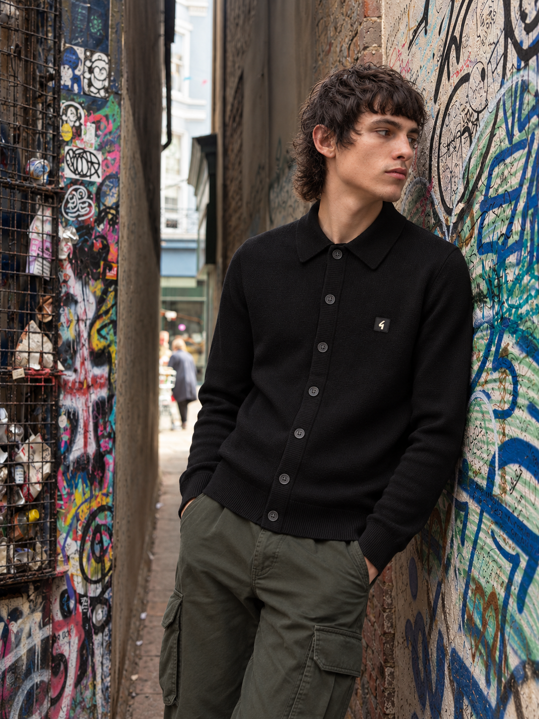 A man wearing a black knit polo cardigan and olive green cargo pants stands in a narrow urban alleyway with graffiti-covered walls.