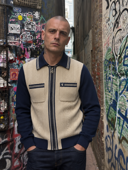 A man wearing a beige and navy blue zip-up knit sweater vest with chest pockets, standing in an urban alleyway with graffiti-covered walls.
