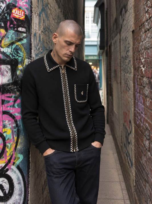 A man wearing a black knit zip-up sweater with gold chain-stitched trim and a chest pocket stands in a narrow urban alleyway with graffiti-covered walls.