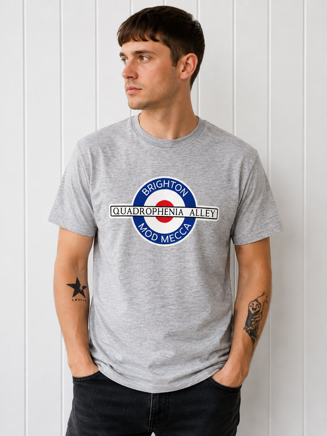 A man wearing a heather gray t-shirt with a circular graphic that reads 'BRIGHTON QUADROPHENIA ALLEY MOD MECCA' around a red, white, and blue target design, standing against a white paneled wall.
