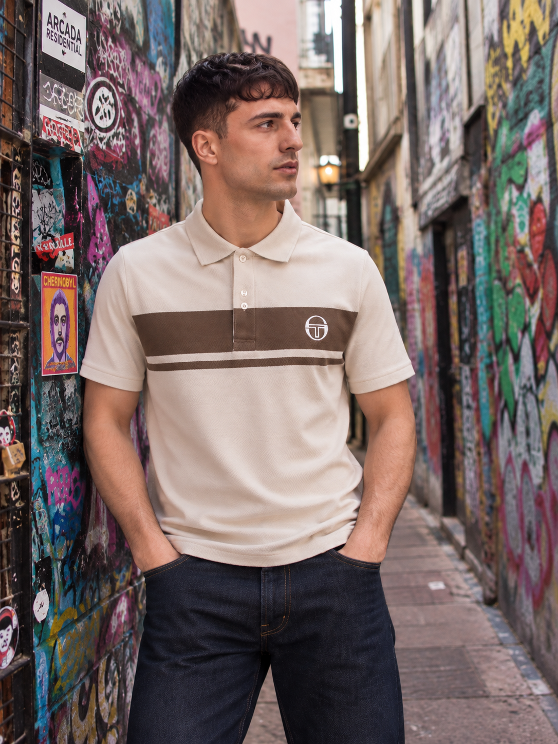 A man wearing a beige short-sleeve polo shirt with a brown chest stripe and a circular logo on the left chest, standing in a narrow urban alleyway with graffiti-covered walls.