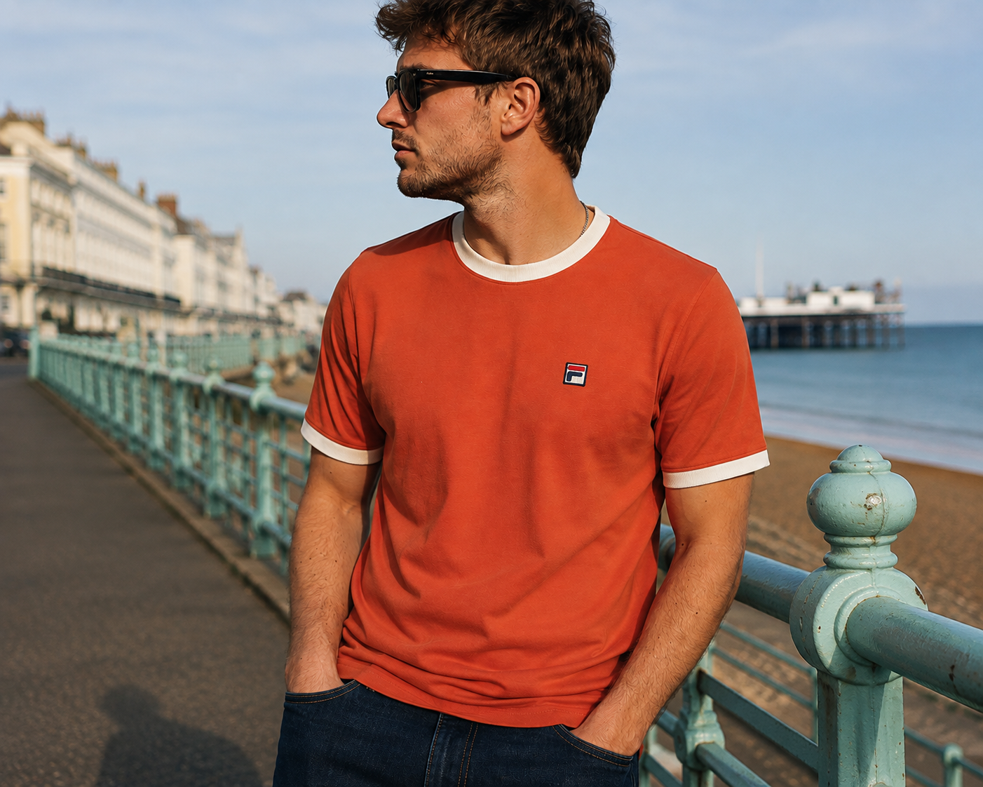 A man wearing a rust-orange Fila ringer t-shirt with white trim, standing on a seaside promenade with a pier and ocean in the background.