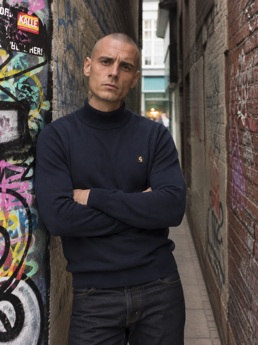 A man with a shaved head stands in a narrow urban alleyway with graffiti-covered walls, wearing a dark navy turtleneck sweater with a small gold embroidered logo on the chest and dark jeans.