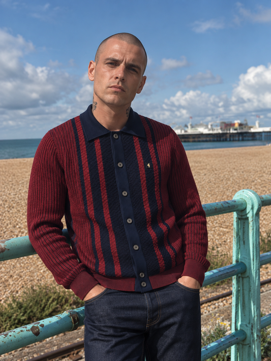 A man wearing a burgundy and navy blue striped knit polo cardigan sweater, standing on a pebble beach with a turquoise railing and a pier in the background.