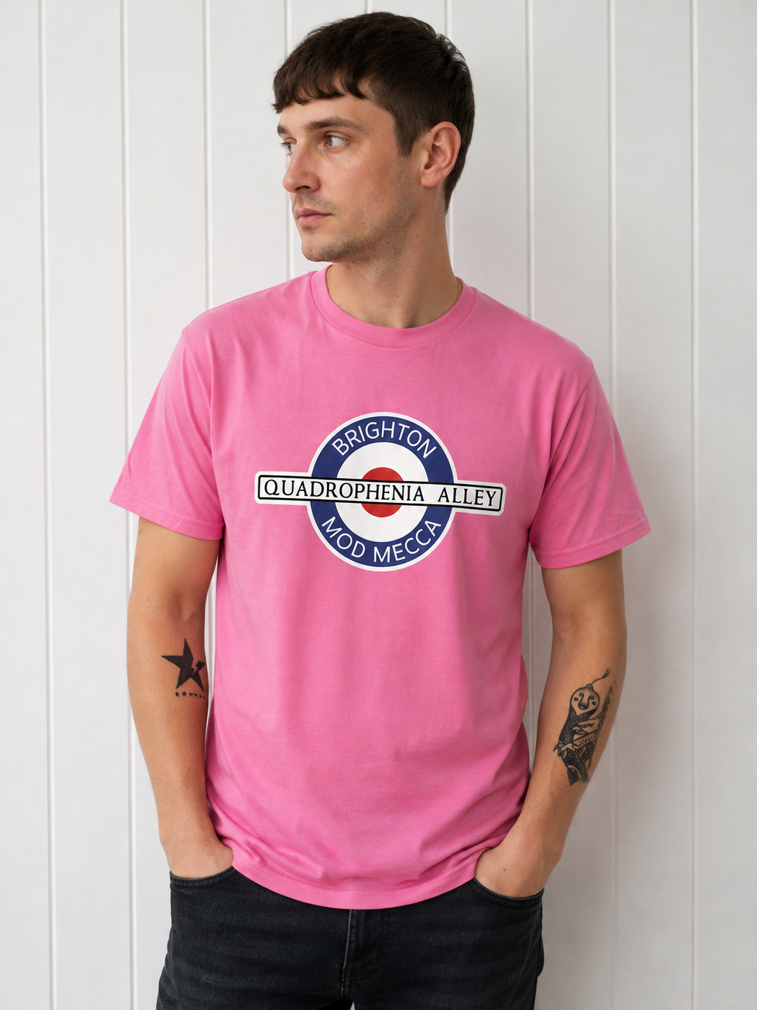 A man wearing a bright pink t-shirt with a circular graphic that reads 'BRIGHTON QUADROPHENIA ALLEY MOD MECCA' in blue, white, and red, standing against a white paneled wall.