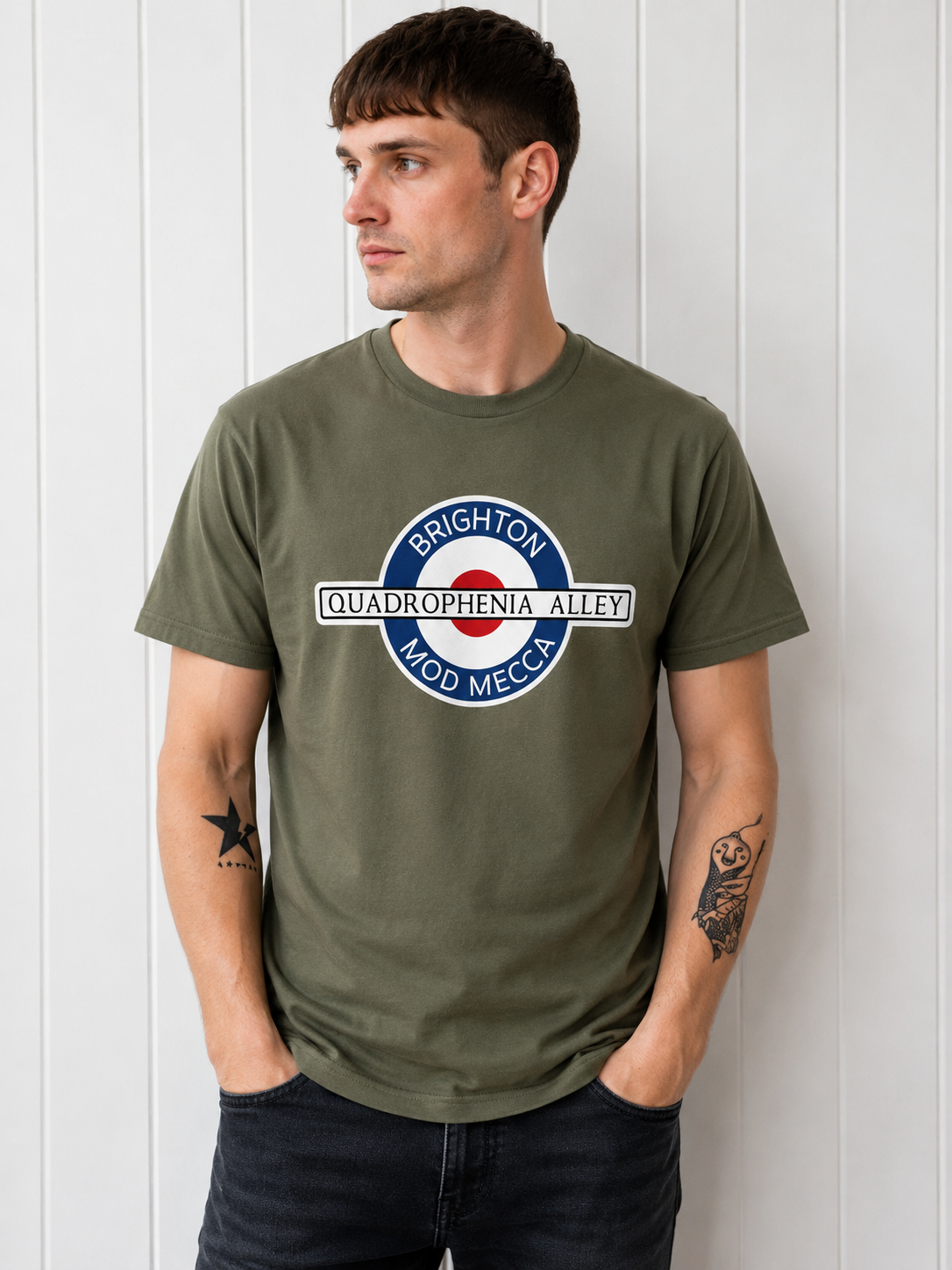 A man wearing an olive green t-shirt with a circular graphic that reads 'BRIGHTON QUADROPHENIA ALLEY MOD MECCA' in blue, white, and red, standing against a white paneled wall.