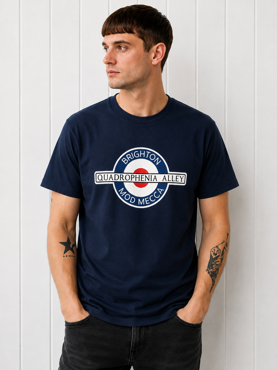 A man wearing a navy blue t-shirt with a circular graphic print that reads 'BRIGHTON QUADROPHENIA ALLEY MOD MECCA' around a red, white, and blue target design, standing against a white paneled wall.