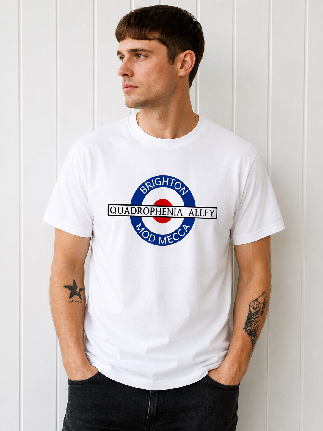 A man wearing a white short-sleeve t-shirt with a circular graphic on the chest that reads 'BRIGHTON QUADROPHENIA ALLEY MOD MECCA' in blue, red, and black, posed against a white paneled wall.
